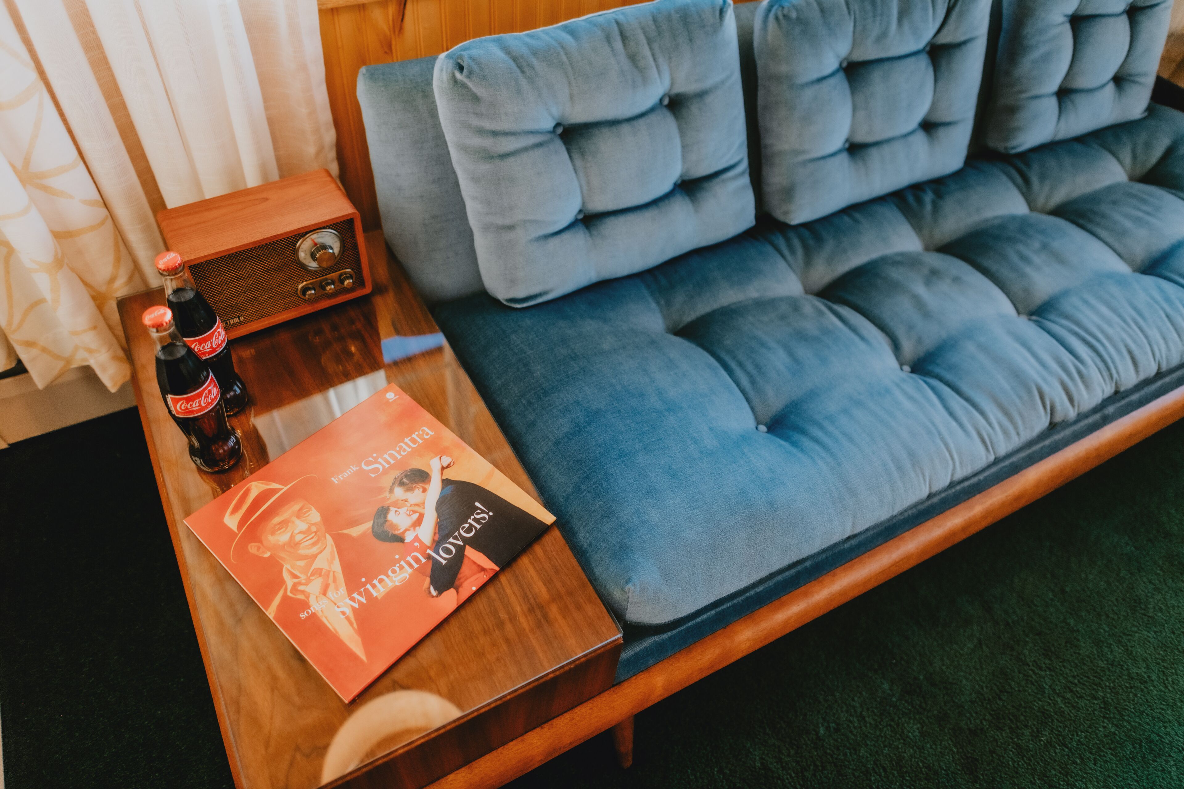 Una habitación con un sofá azul con capitoné, una mesita de madera con una botella de Coca-Cola y una revista.