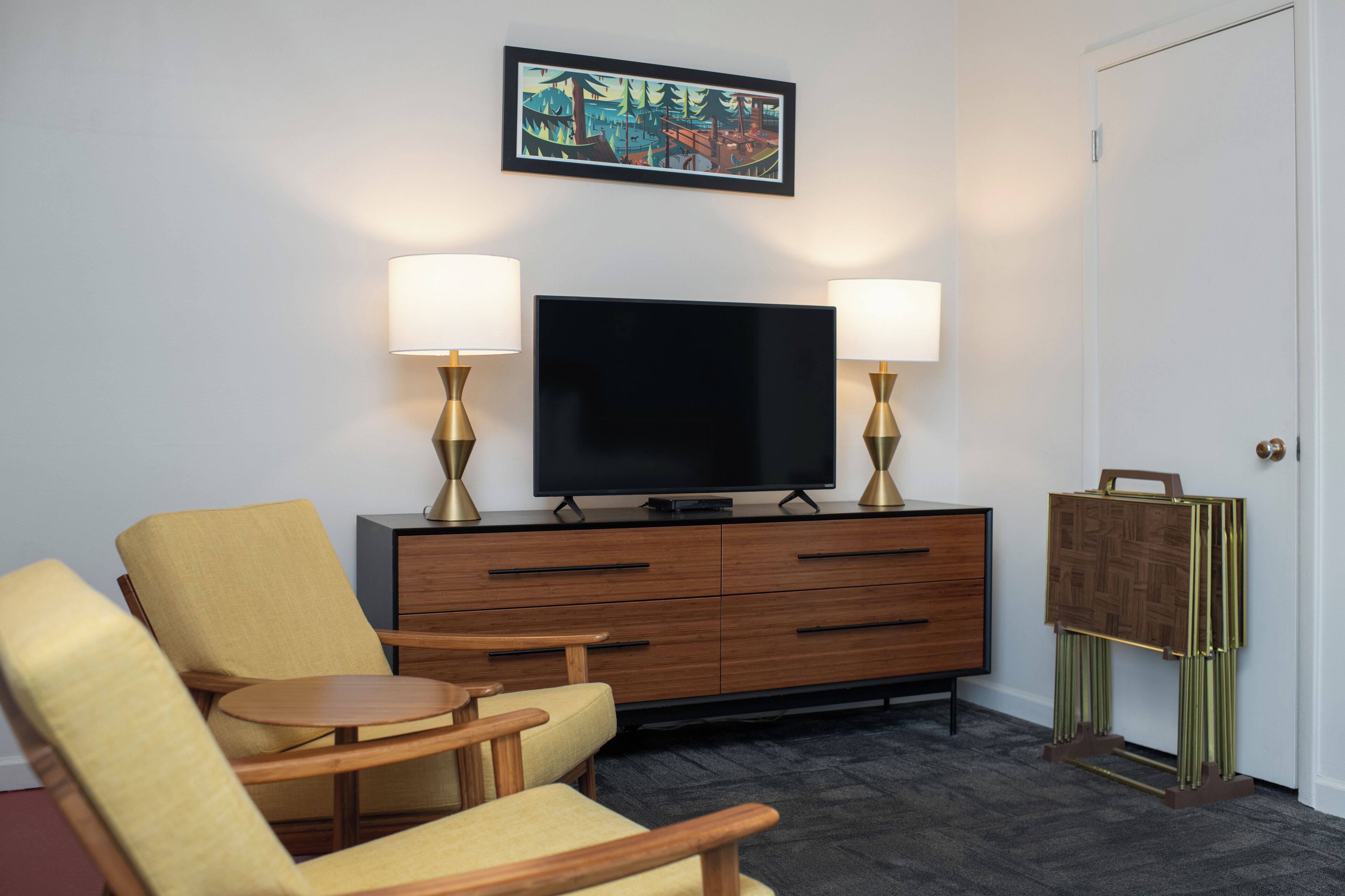 Hotelkamer met een flatscreen televisie, twee lampen, een houten kast, een stoel, een klein tafeltje en een deur.