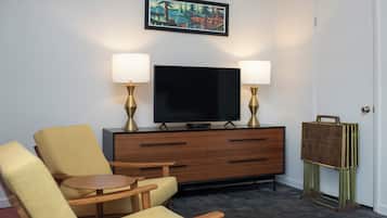 A hotel room with a flat-screen TV, two lamps, a wooden cabinet, a chair, a small table, and a door.