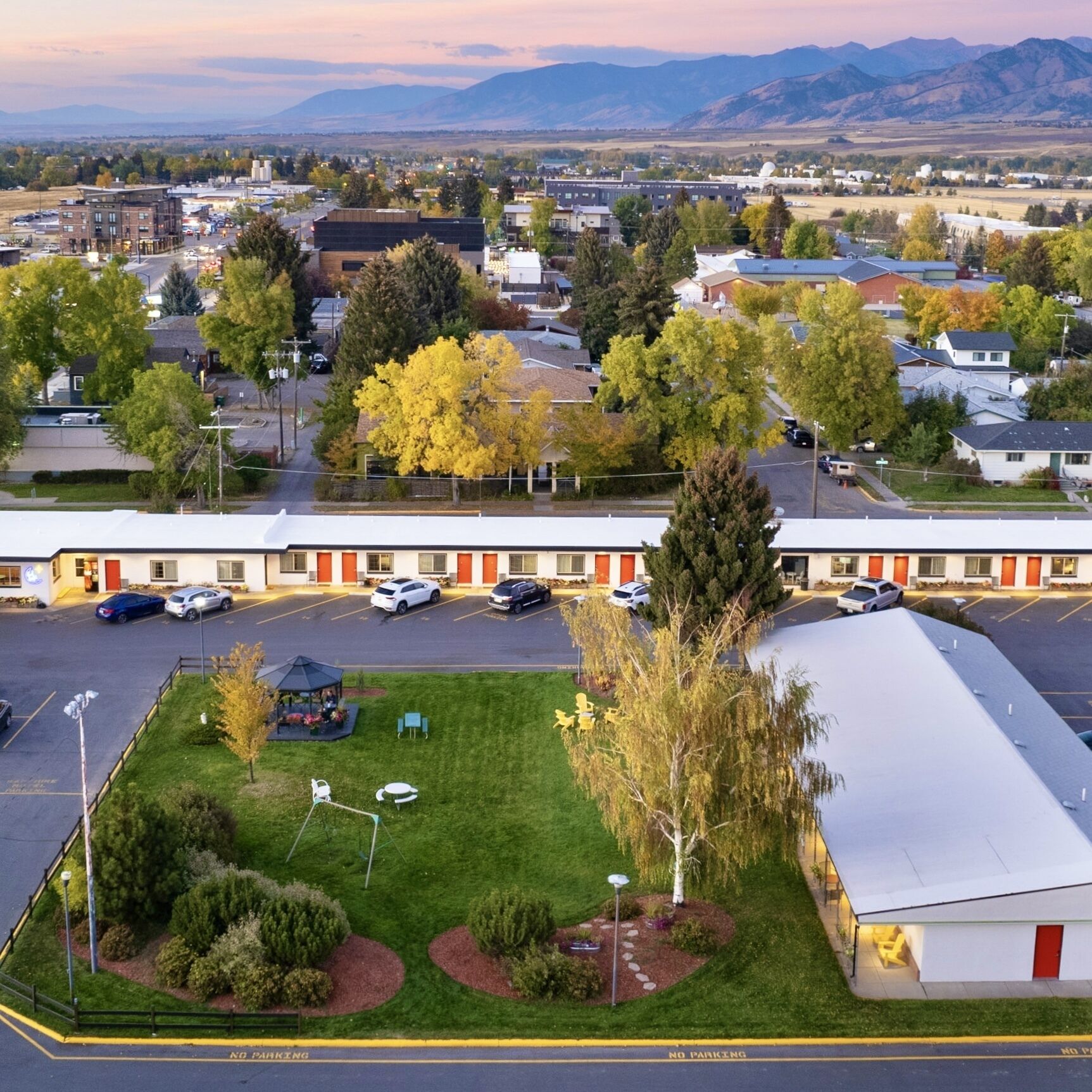 Un motel doté d’un parking, d’une petite pelouse avec des sièges extérieurs, et offrant une vue sur une ville et des montagnes en arrière-plan.