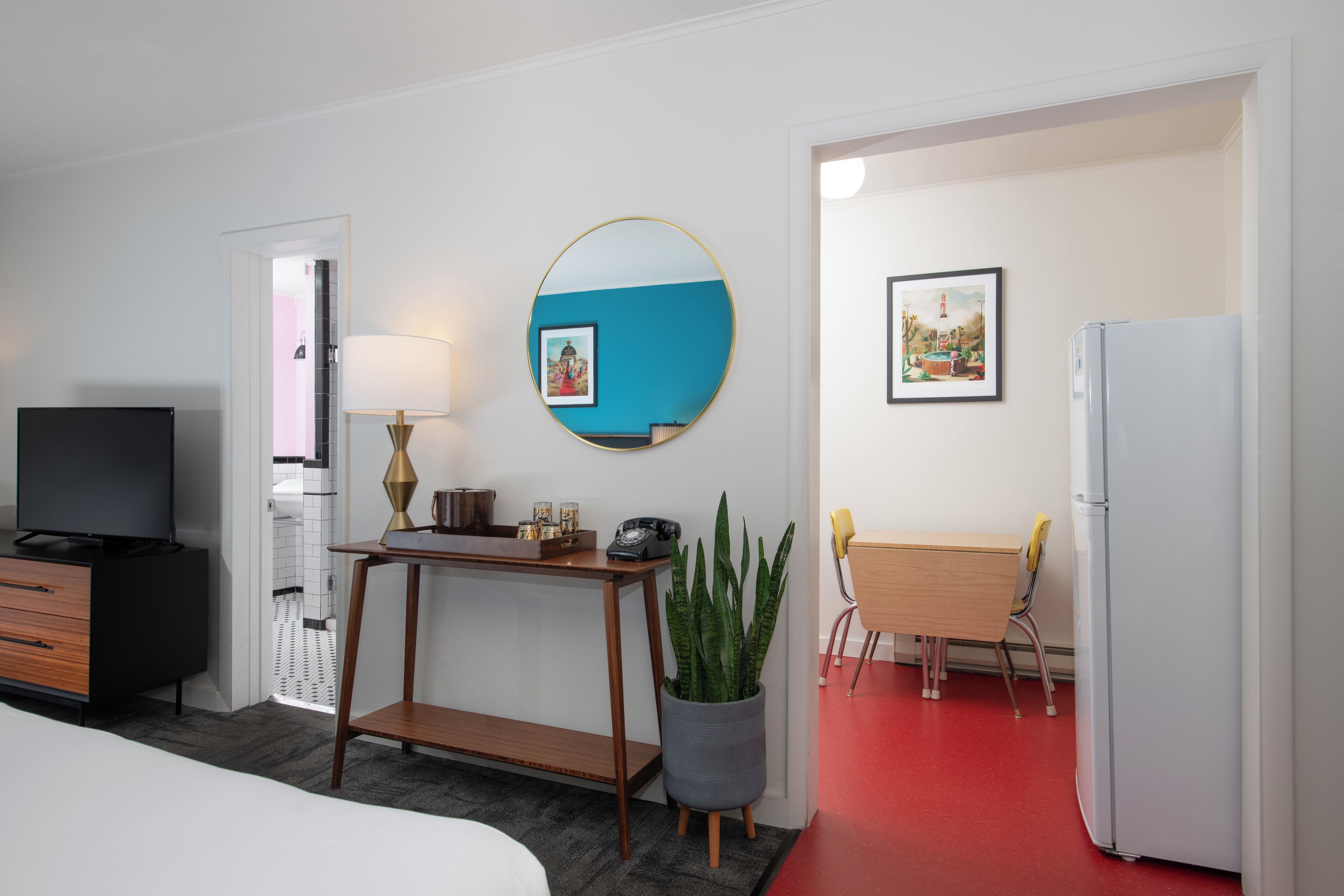 A hotel room with a bed, a desk with a lamp, a television, a potted plant, a dining table with chairs, and a refrigerator.