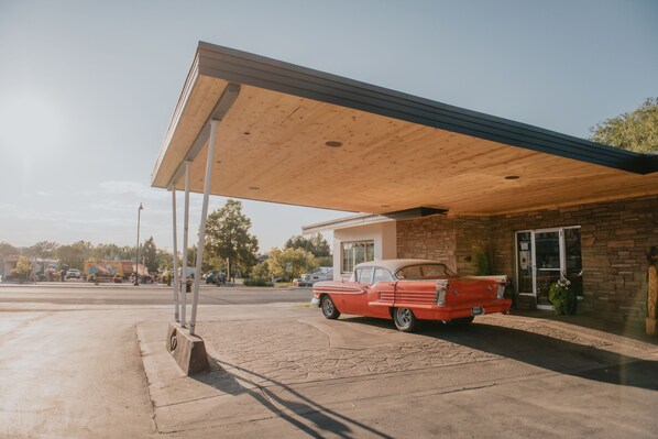 A vintage red car parked under a covered area with a brick building in the background.