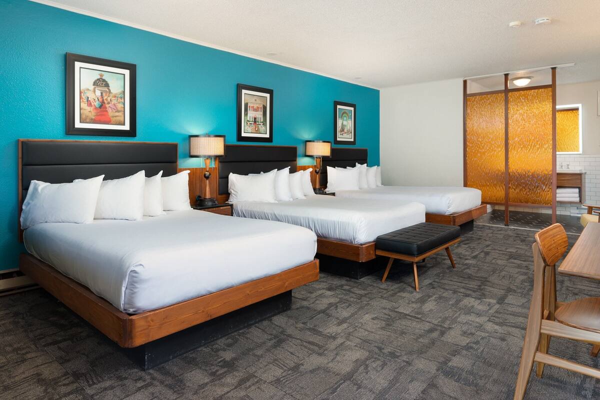 A hotel room with two beds, a wooden headboard, a bench, a chair, and a bathroom with a tiled wall and a sink.