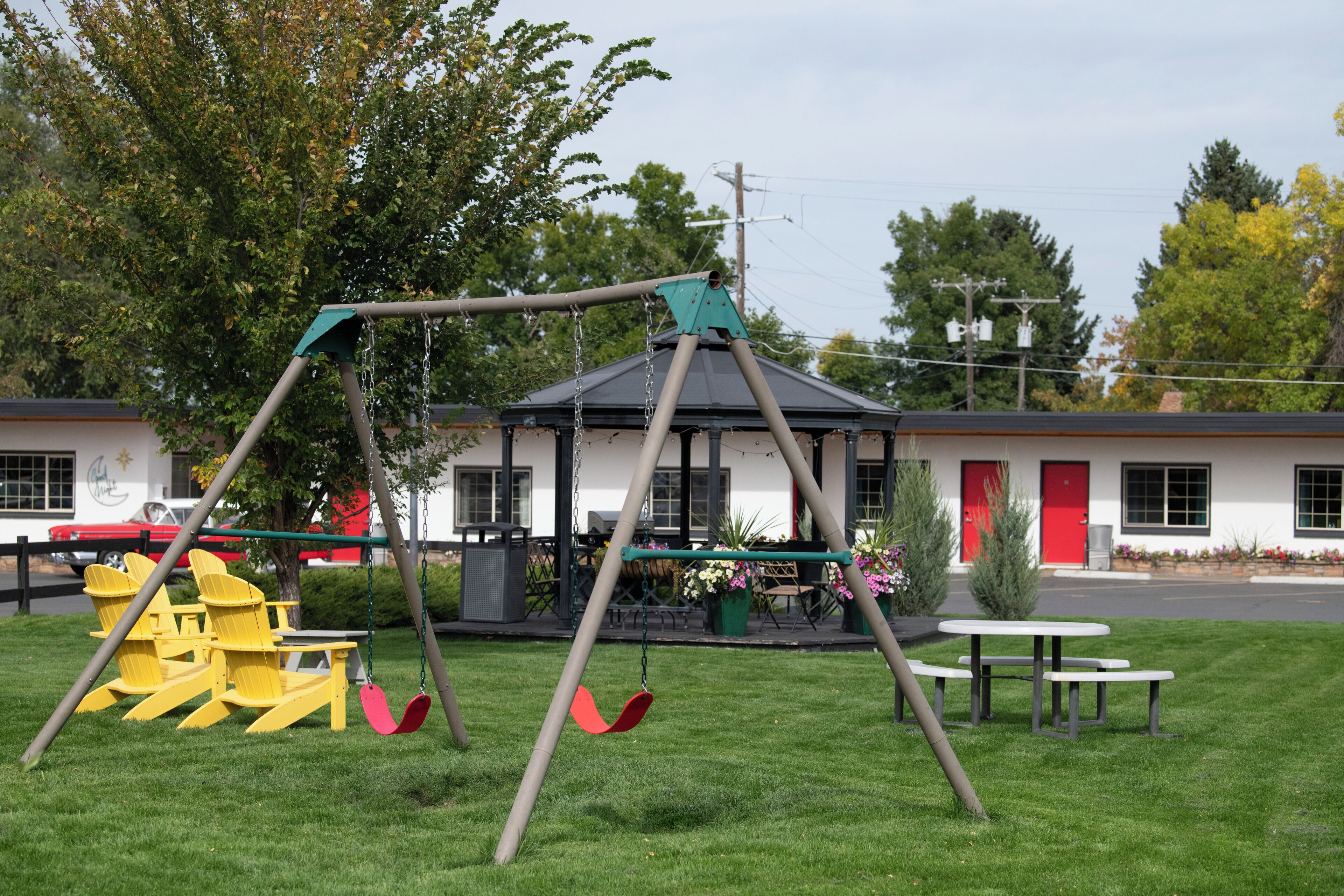 Een speeltuin met schommels en stoelen, een picknicktafel en een gebouw met een rode deur op de achtergrond.