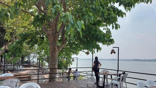 Una terraza al aire libre con piso de madera, sillas y mesas de plástico blanco. Una persona está de pie cerca de la barandilla, mirando un cuerpo de agua.