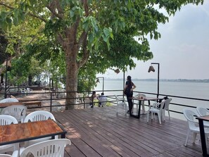 An outdoor deck with wooden flooring, white plastic chairs, and tables. A person is standing near the railing overlooking a body of water.
