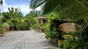 A paved pathway leading to a building surrounded by lush greenery and a signboard with contact information. - Anse Kerlan Beach Chalets (Praslin Island)