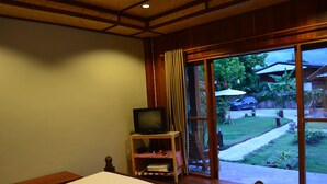 A bedroom with a bed, a television on a wooden stand, and a view of a garden through a sliding glass door. - Baan Aew Pai (Pai)