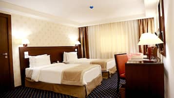 Quarto de hotel com duas camas, uma escrivaninha, uma cadeira e uma luminária de mesa.