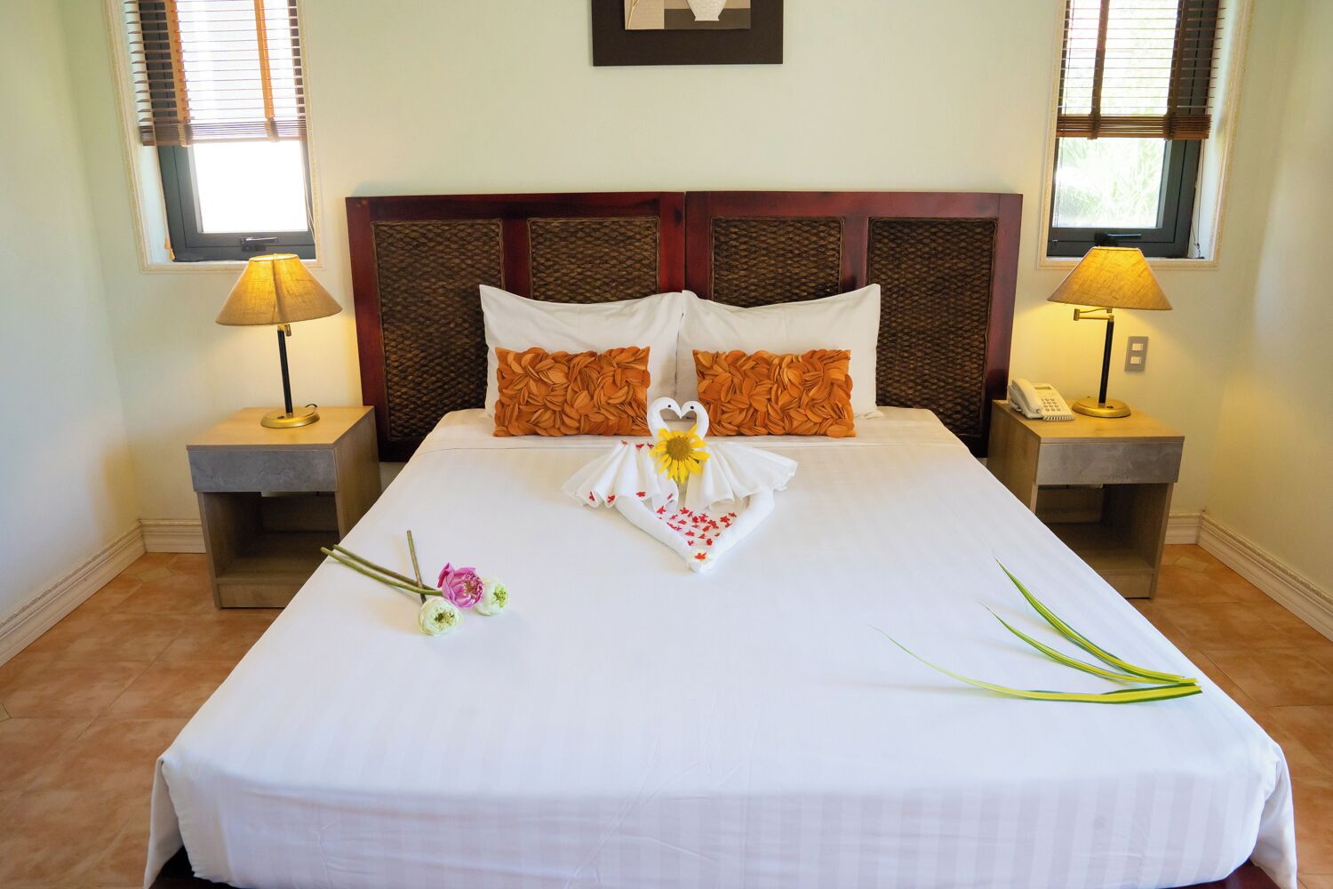 Una cama bien hecha con una colcha blanca, dos almohadas decorativas, un arreglo de toallas con forma de corazón y un pequeño ramo de flores.