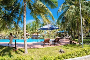 Un área junto a la piscina con sillones de descanso, sombrillas y palmeras.