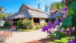 A traditional thatched-roof building with a wooden porch, surrounded by lush greenery and flowering plants. - Lotus Vung Tau Resort (Ho Chi Minh City)