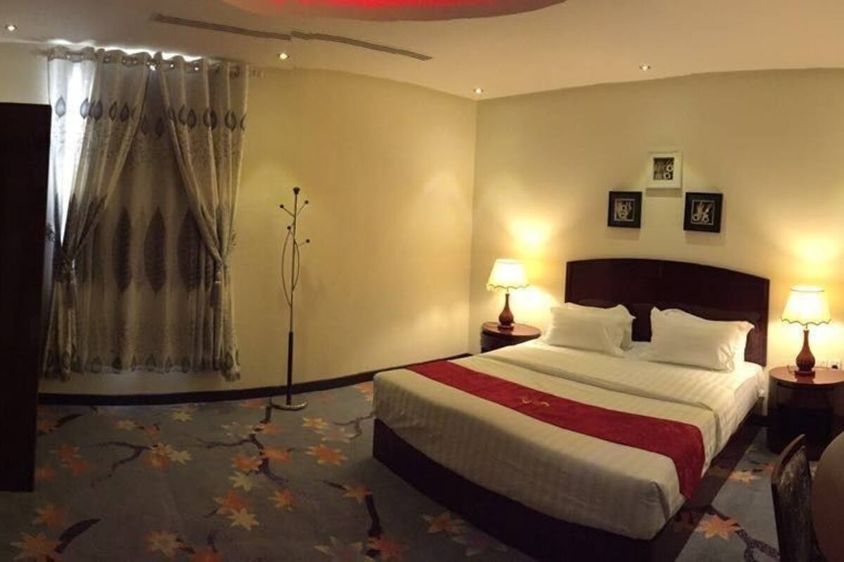 Ein Hotelzimmer mit Bett, zwei Nachttischlampen, einem Flachbildfernseher, einem Kleiderhaken und einem Fenster mit Vorhängen.