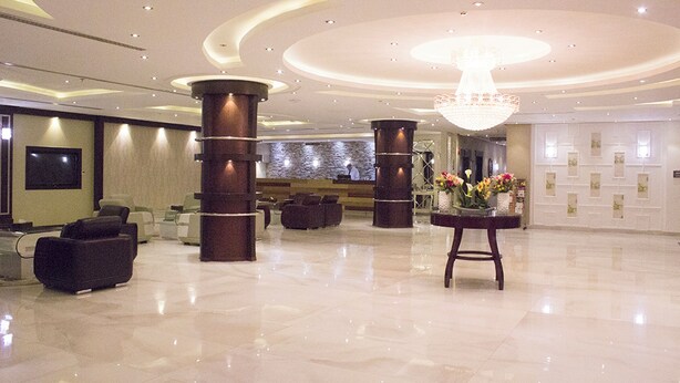 A spacious lobby with a high ceiling, large columns, and a central chandelier. There are several seating areas and a reception desk with a vase of flowers. - Merfal Serviced Apartments-Royal (Riyadh)