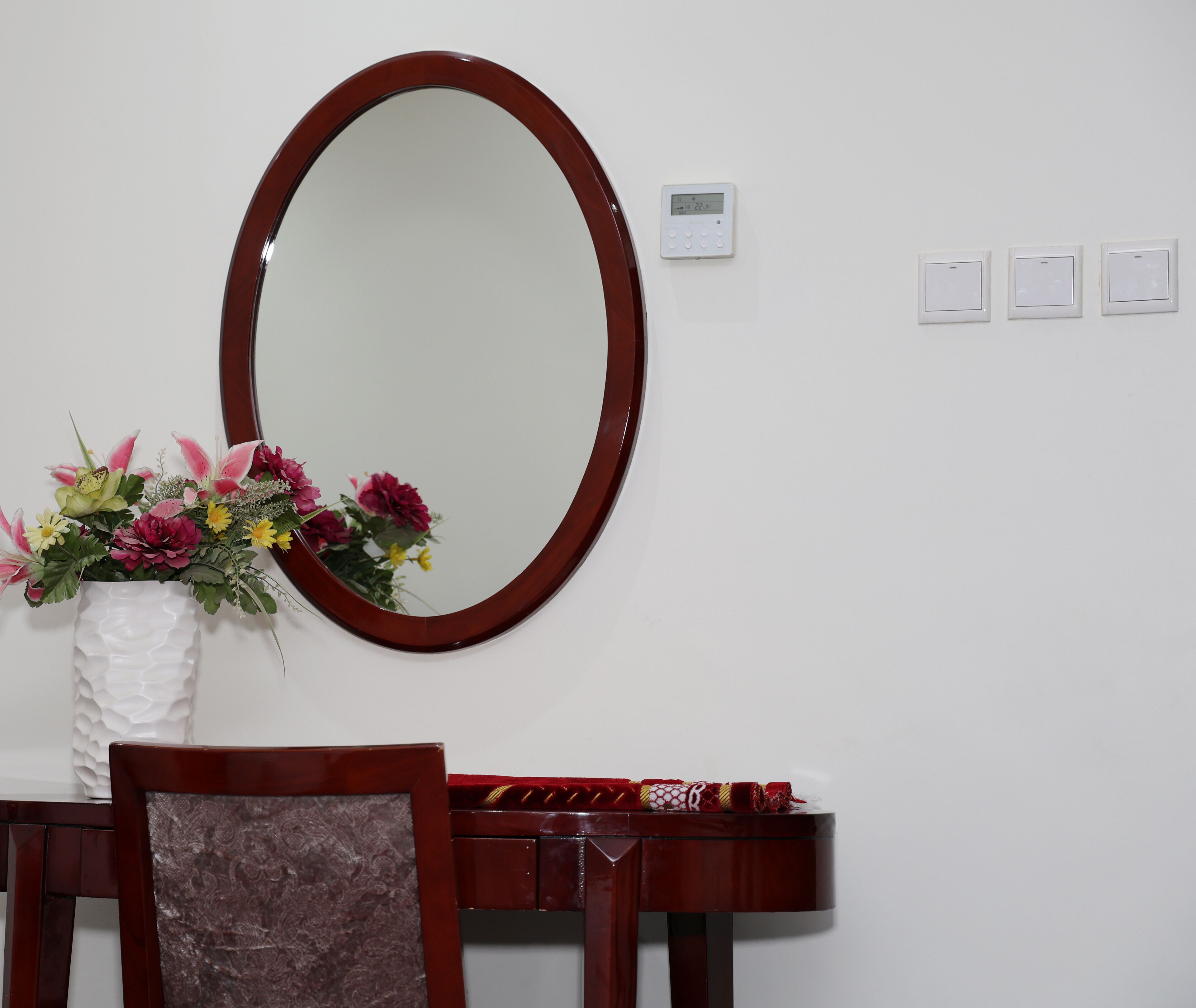 Una habitación con un espejo ovalado grande, un florero blanco con flores, una silla de madera y una mesita con un mantel rojo.
