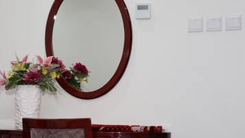Una habitación con un espejo ovalado grande, un florero blanco con flores, una silla de madera y una mesita con un mantel rojo.
