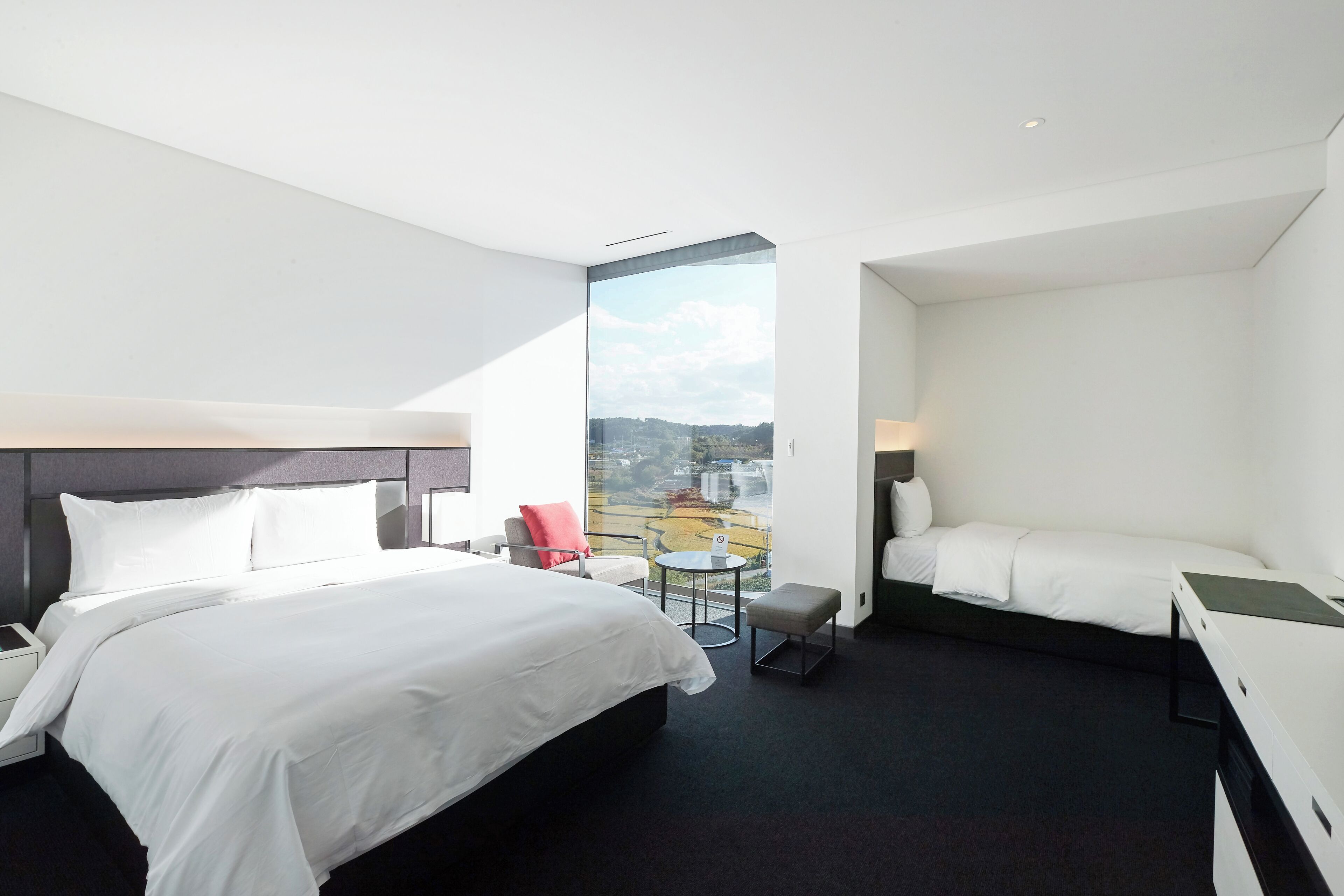 A modern hotel room with a large bed, a smaller bed, a desk, and a chair. There is a balcony with a view of the city.