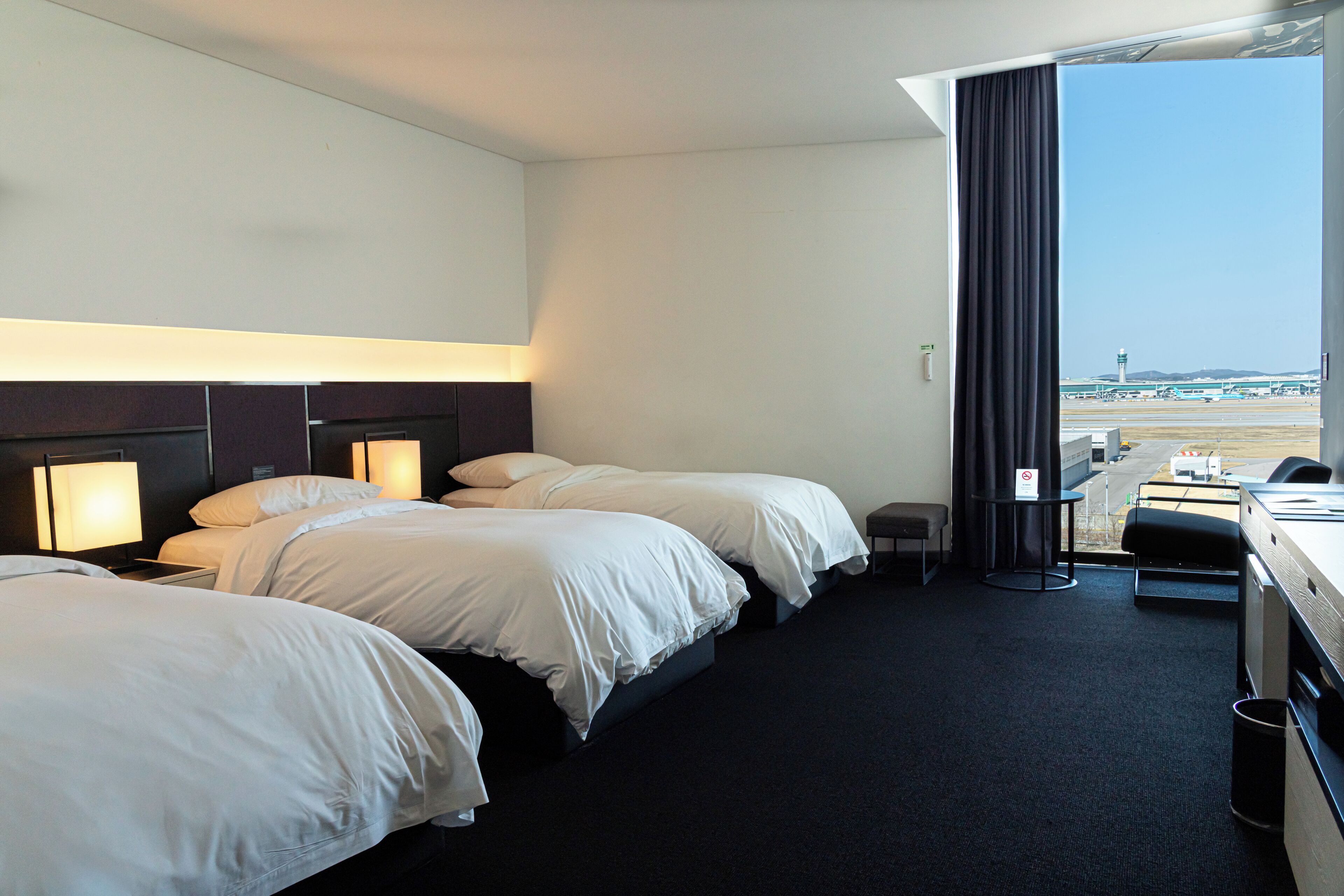 Habitación de hotel con dos camas, un escritorio, una silla y vista al aeropuerto.