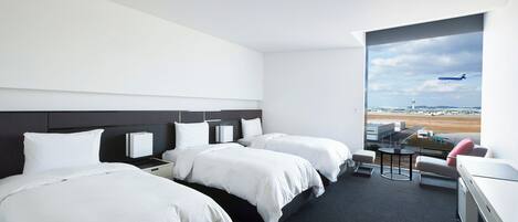 Habitación de hotel con dos camas, un escritorio, una silla y un ventanal con vista al aeropuerto.