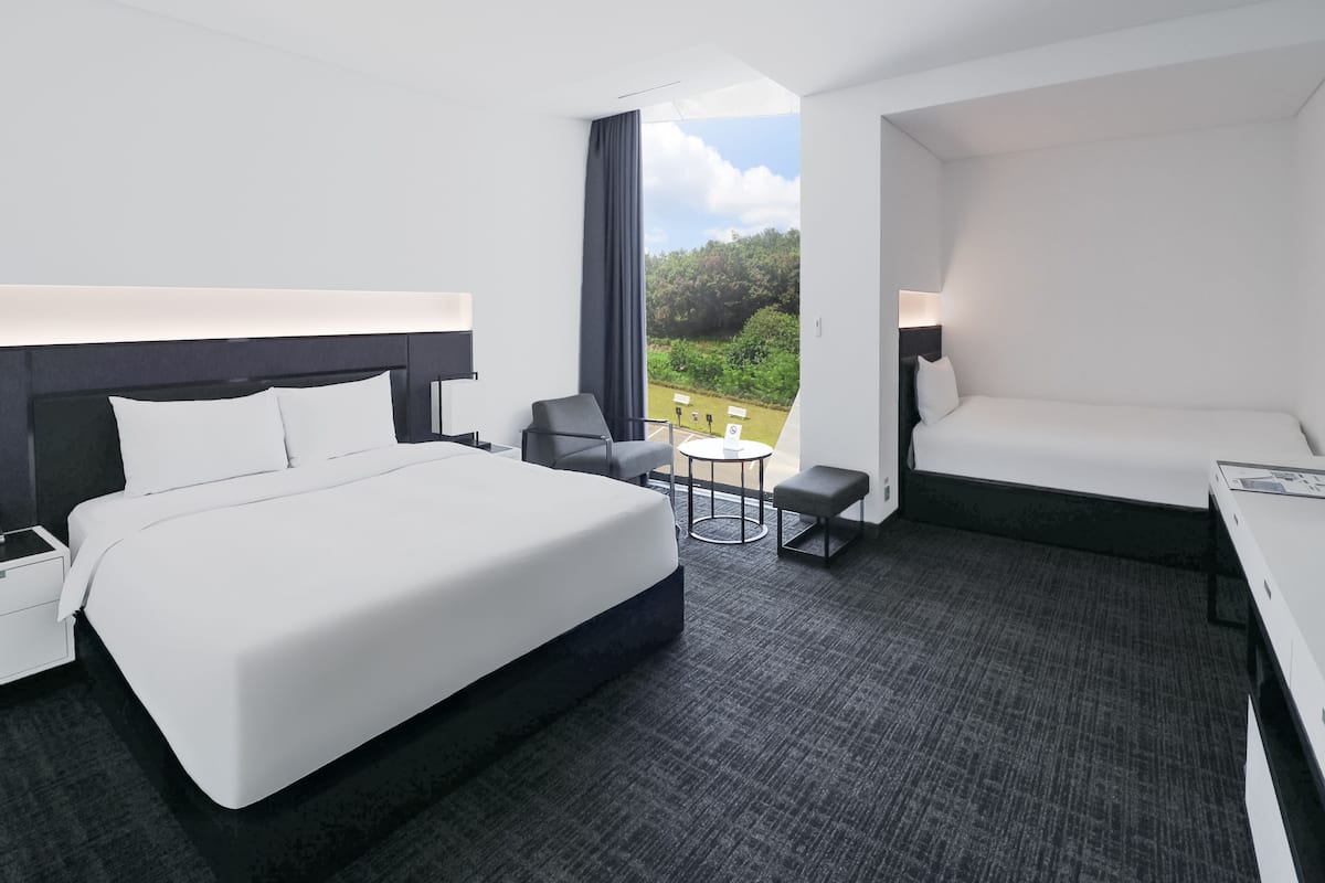 A hotel room with a large bed, a desk, a chair, and a small table. There is a window with a view of greenery outside.