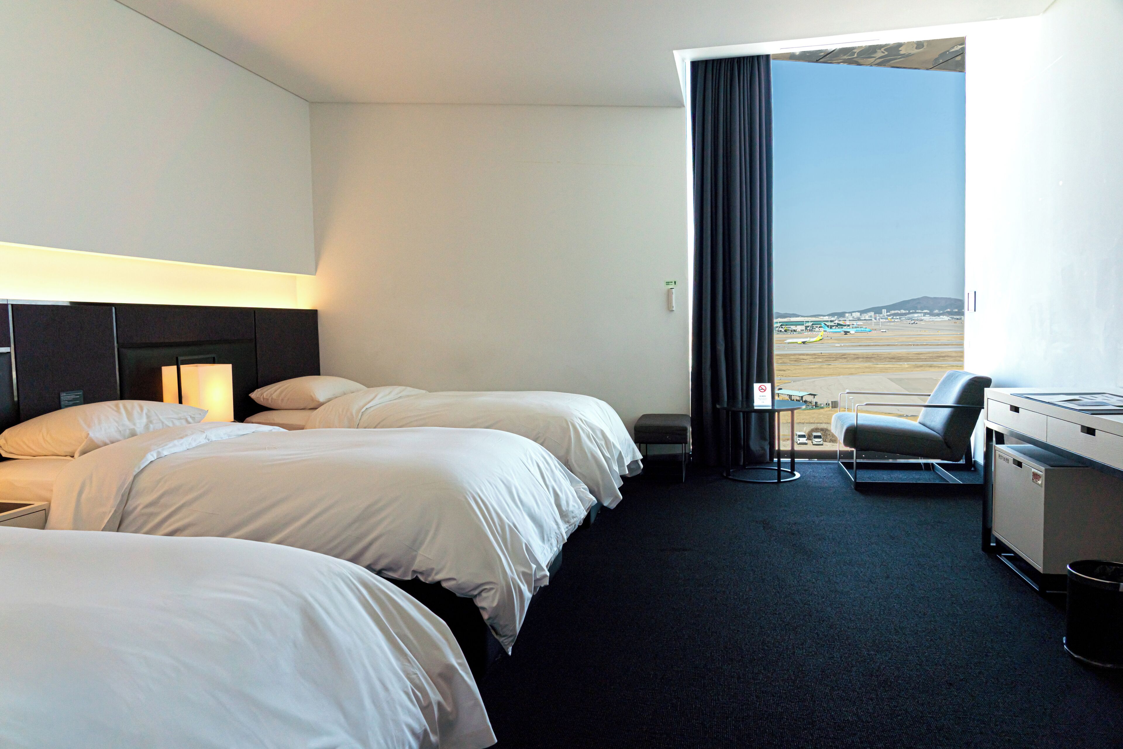 Een hotelkamer met twee bedden, een bureau, een stoel en uitzicht op het vliegveld.