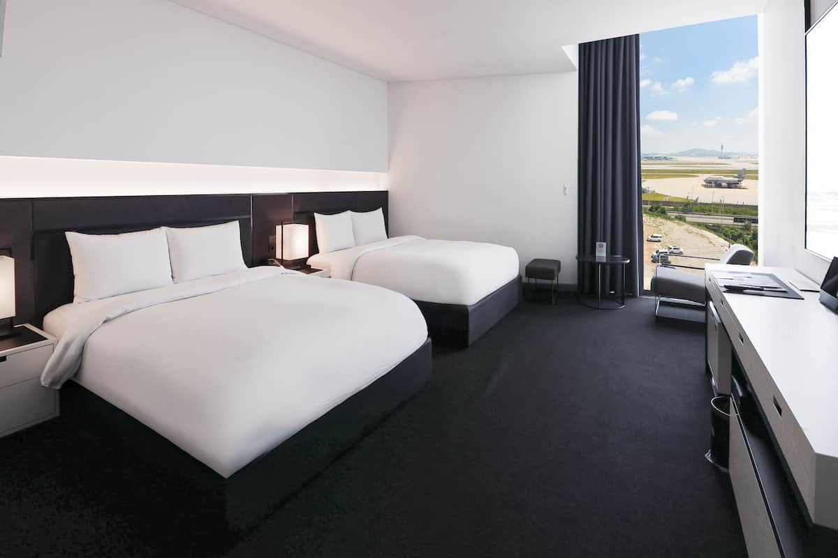 A modern hotel room with two beds, a desk, and a view of the airport.