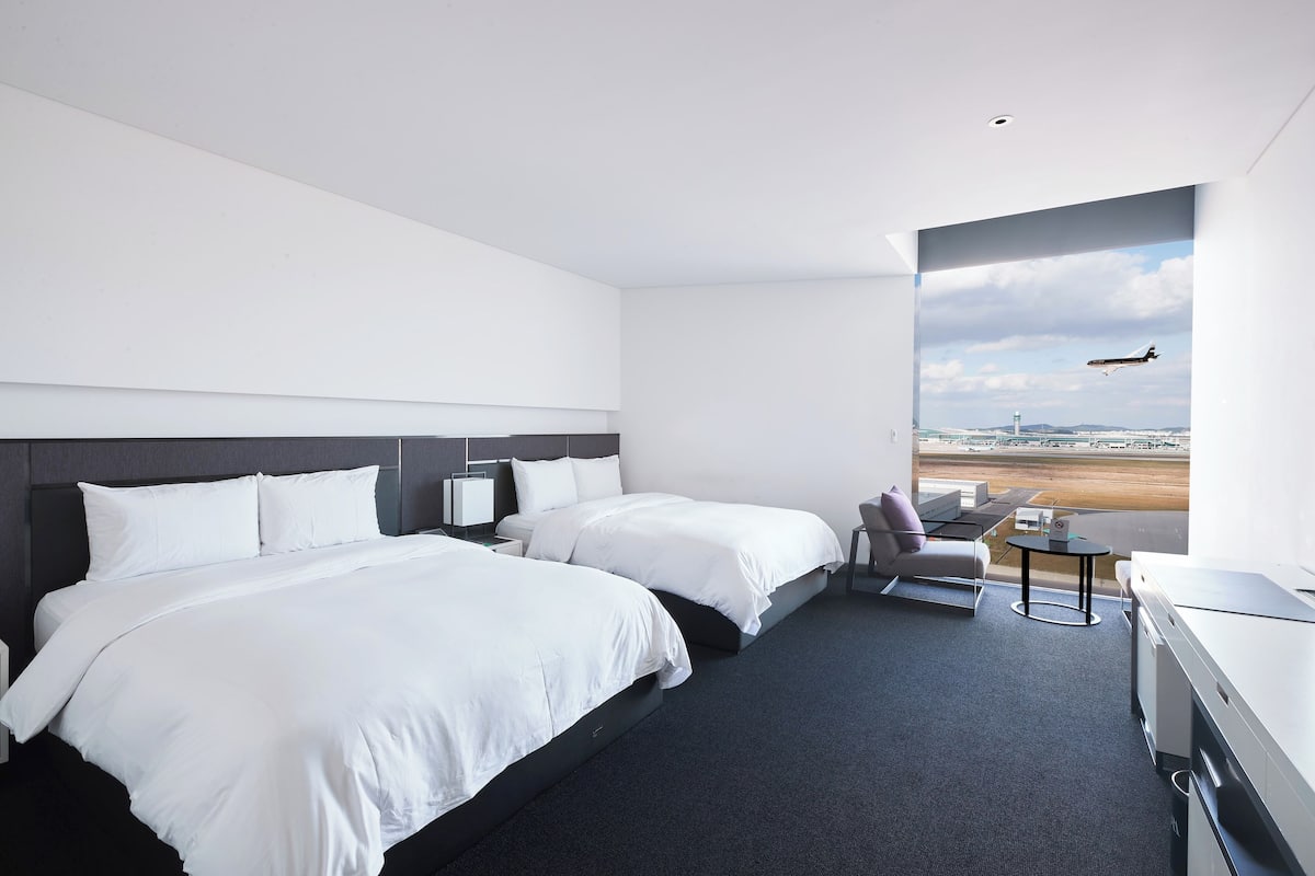 A hotel room with two beds, a desk, a chair, and a view of an airport.