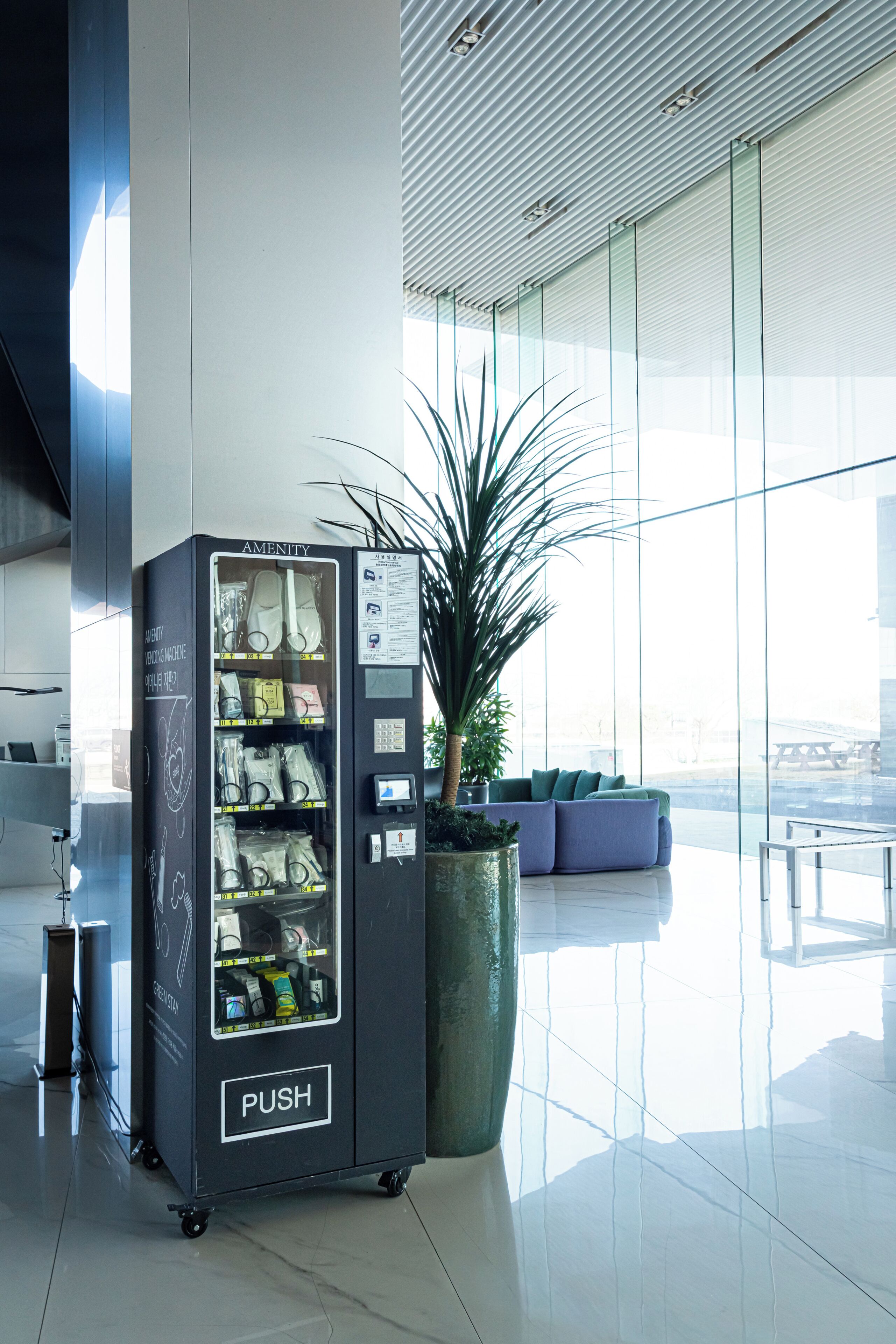 A vending machine labeled 'AMENITY' with a 'PUSH' button, a potted plant, and a seating area with a view.