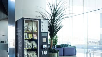 Ein Getränkeautomat mit dem Aufdruck 'AMENITY' und einem 'DRUCKEN'-Knopf, eine Topfpflanze und eine Sitzecke mit Blick.