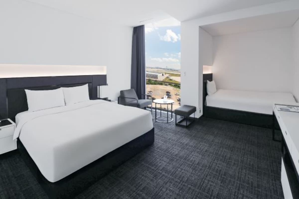 A modern hotel room with a large bed, a desk, a chair, and a small table. There is a view of the outside through a large window.