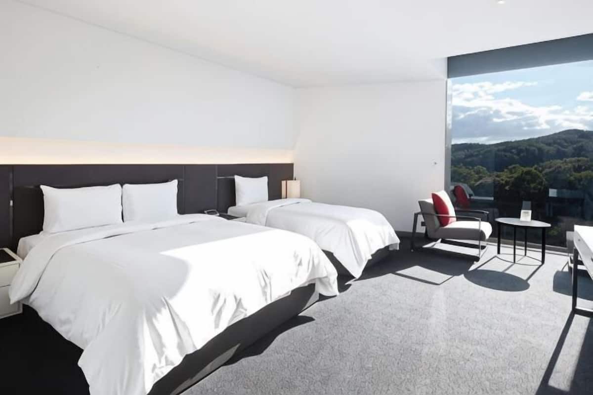 A modern hotel room with two beds, a small table, a chair, and a view of the outdoors.