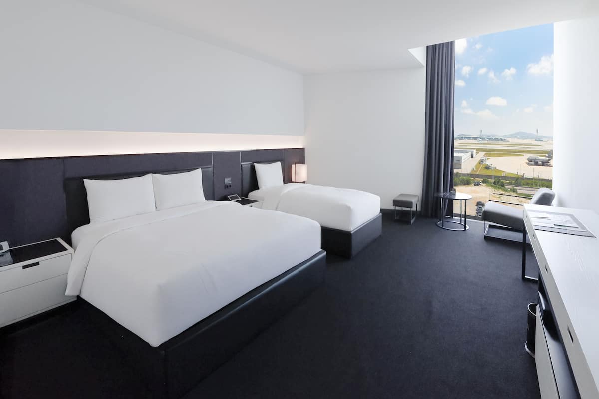 A modern hotel room with two beds, a desk, and a large window with a view of the airport.