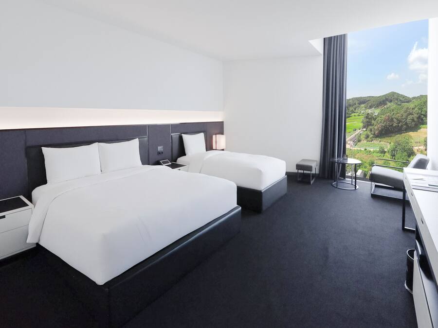 A modern hotel room with two beds, a desk, and a large window offering a view of a green landscape.