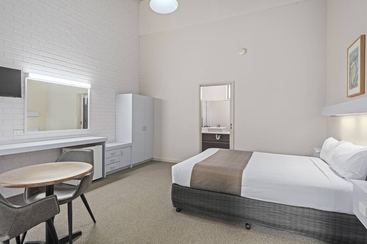 A hotel room with a bed, a desk with a chair, a wardrobe, a mirror, and a light fixture.