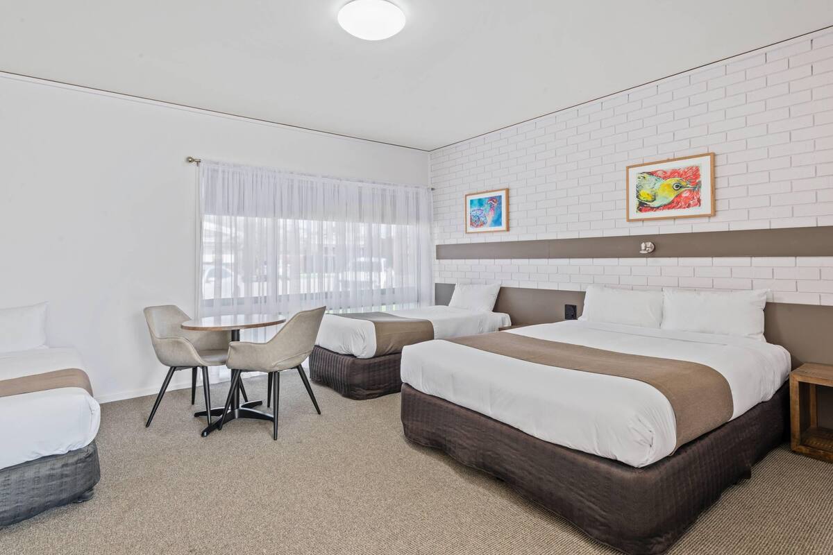 A hotel room with a bed, two chairs, a small table, a window with sheer curtains, and two framed pictures on the wall.