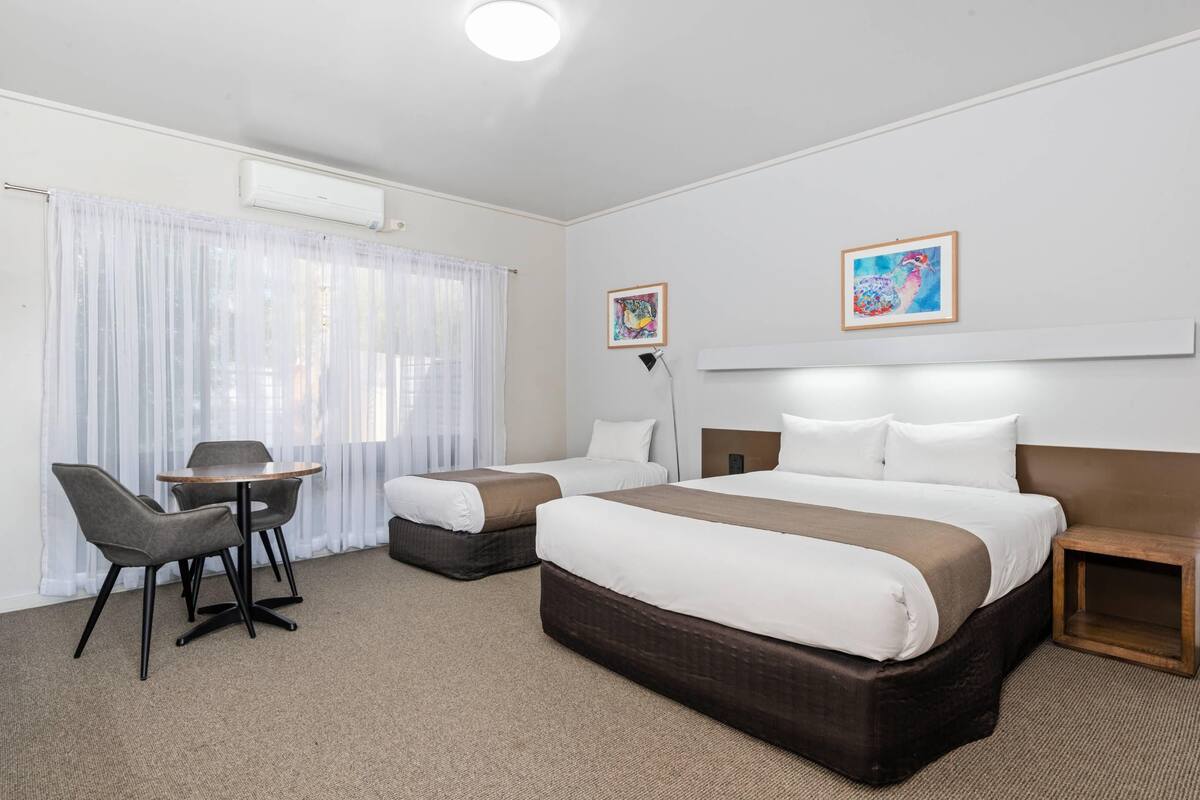 A hotel room with a bed, two bedside tables, a chair, a small table, and a window with sheer curtains.