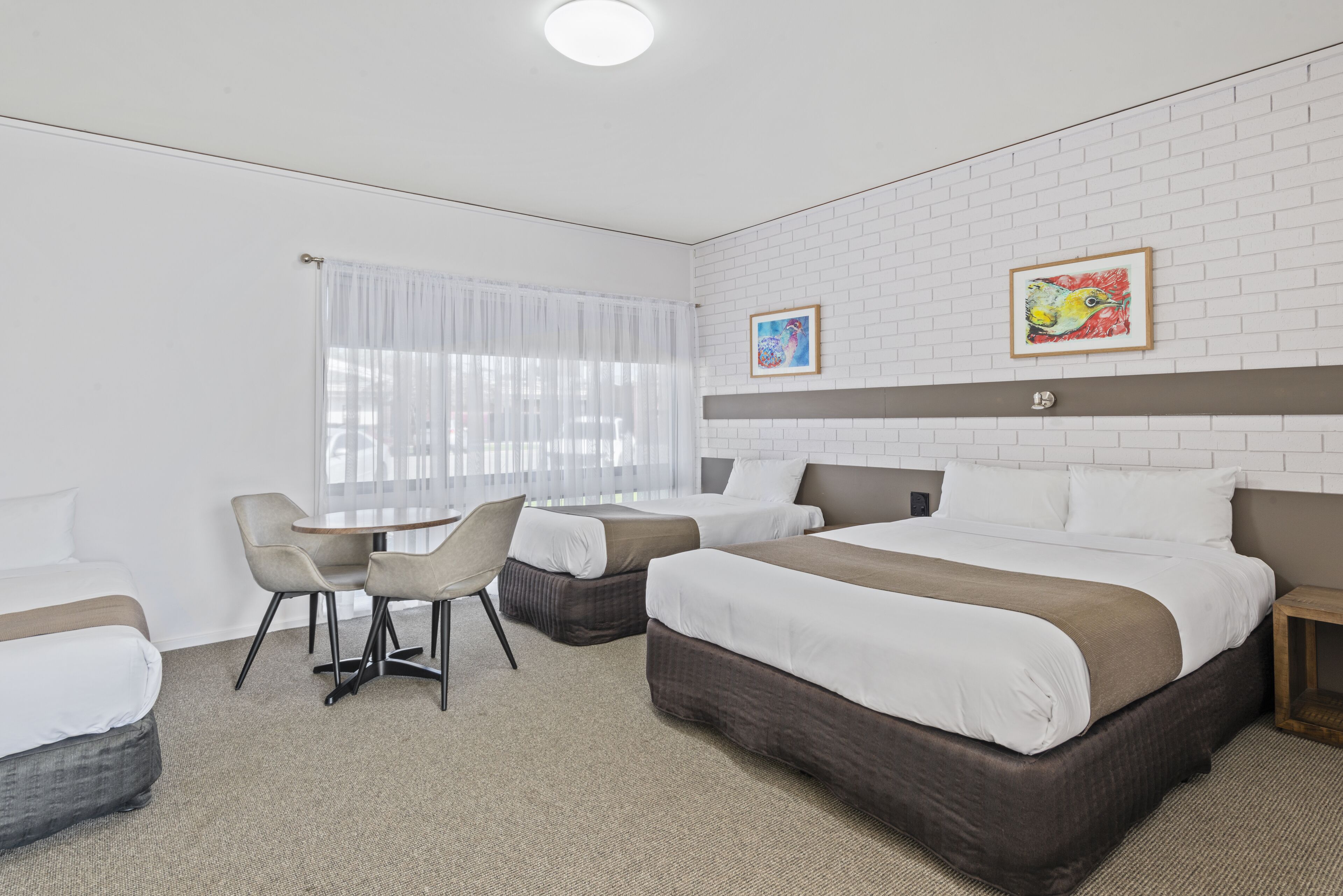 A hotel room with two beds, a small table, and chairs. There is a window with sheer curtains and a painting on the wall.