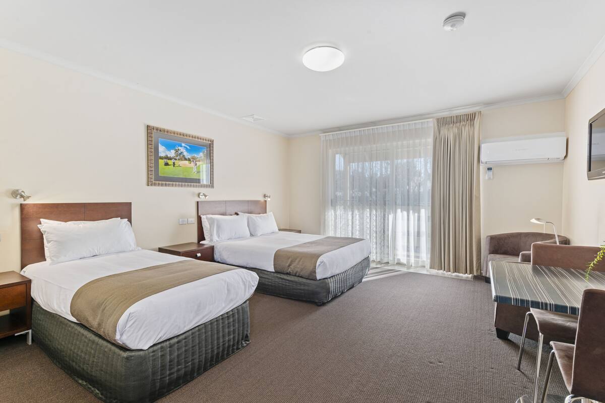 A hotel room with two beds, a desk, and a chair. There is a window with curtains, a picture on the wall, and an air conditioning unit.