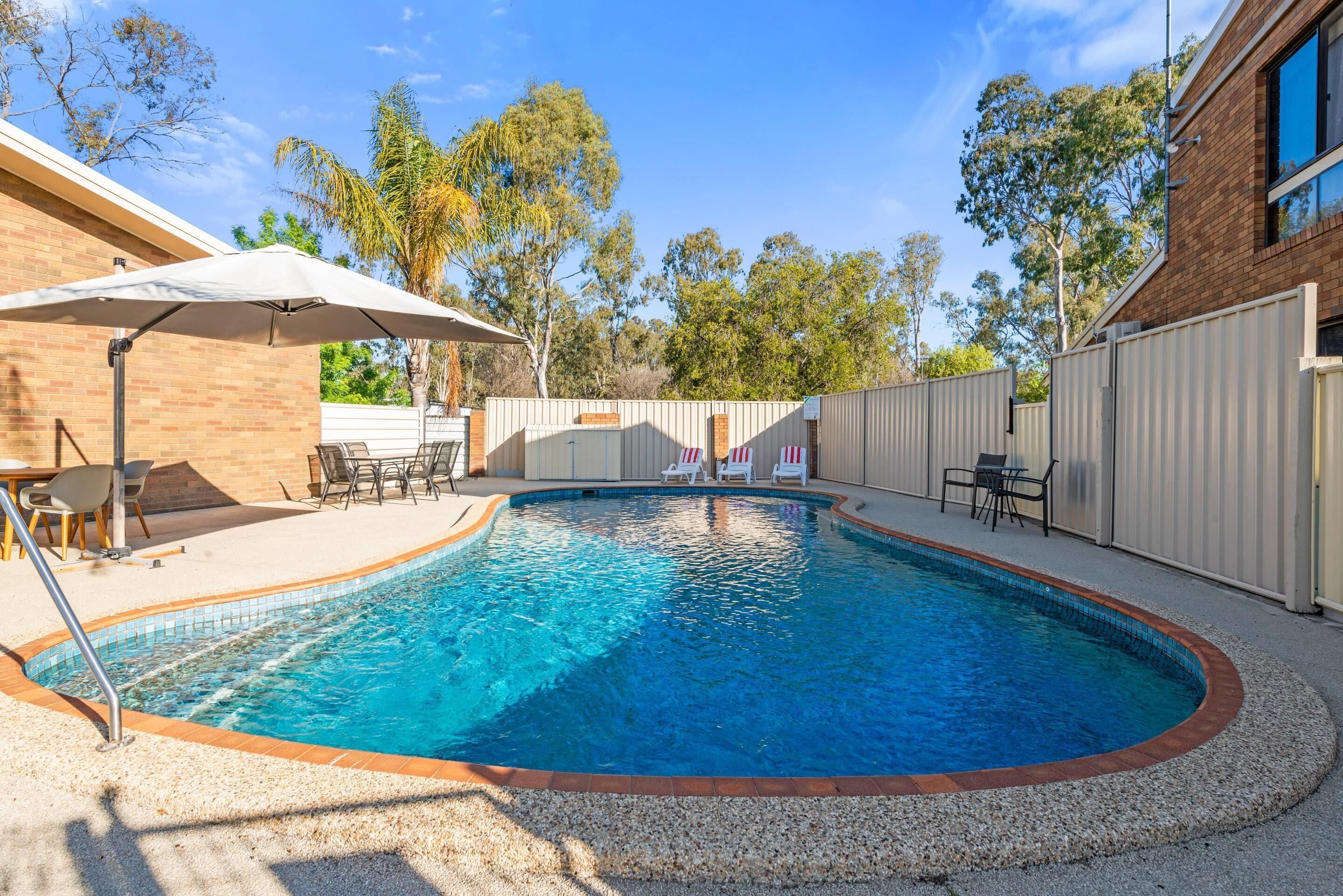 A swimming pool with a tiled edge, surrounded by a gravel area, and a covered patio with chairs and an umbrella.