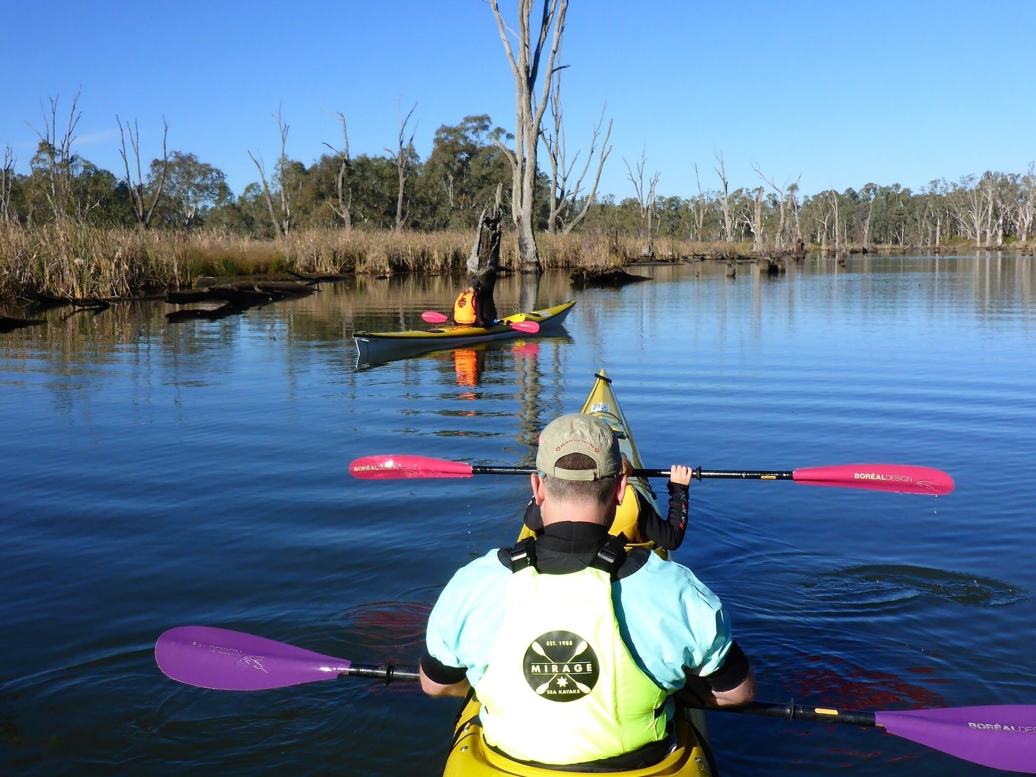 Un kayakista en un kayak amarillo, con chaleco salvavidas y sombrero, remando en un río tranquilo.