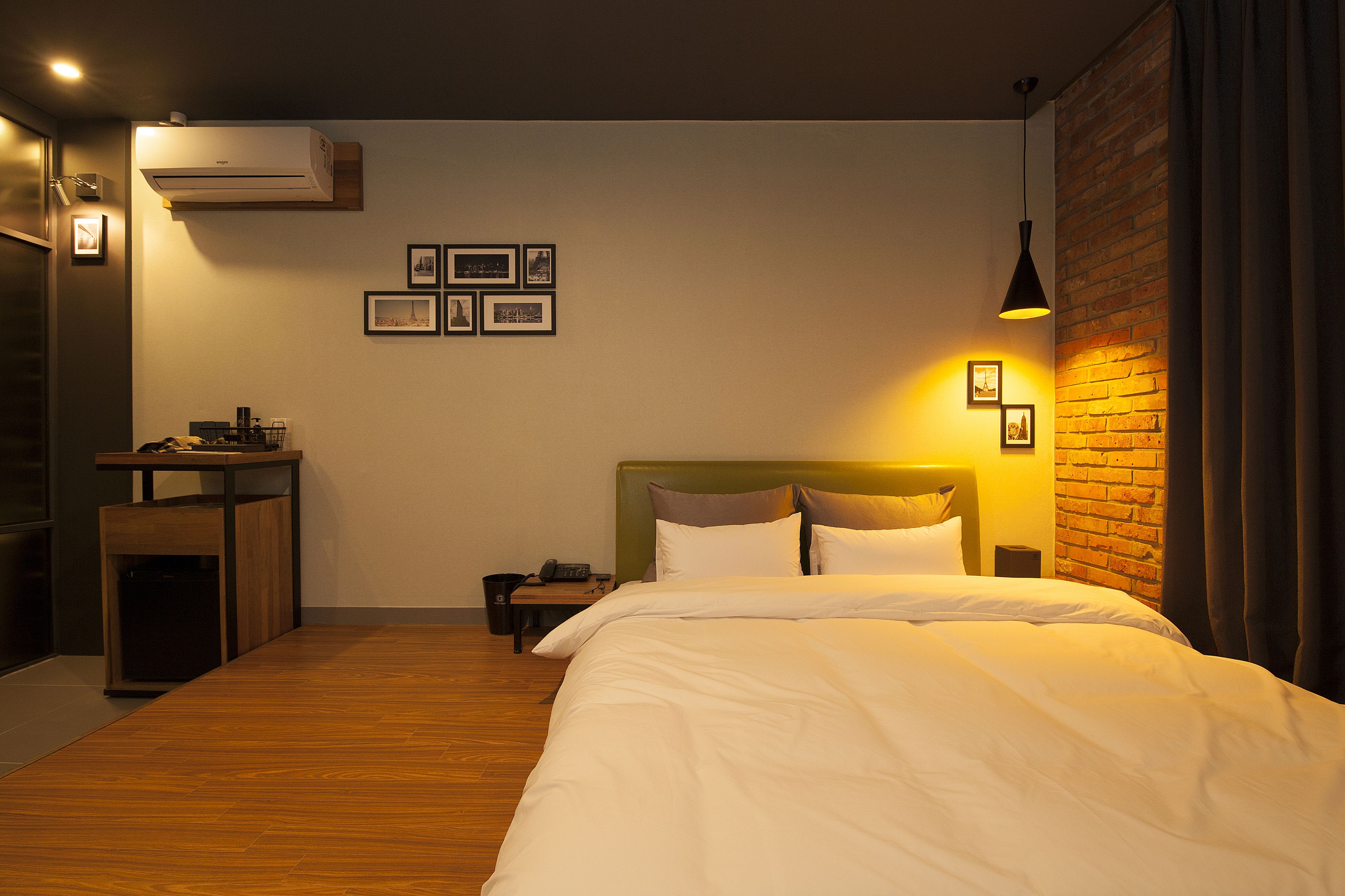 A hotel room with a bed, a small table, a trash bin, and a wall-mounted air conditioner.