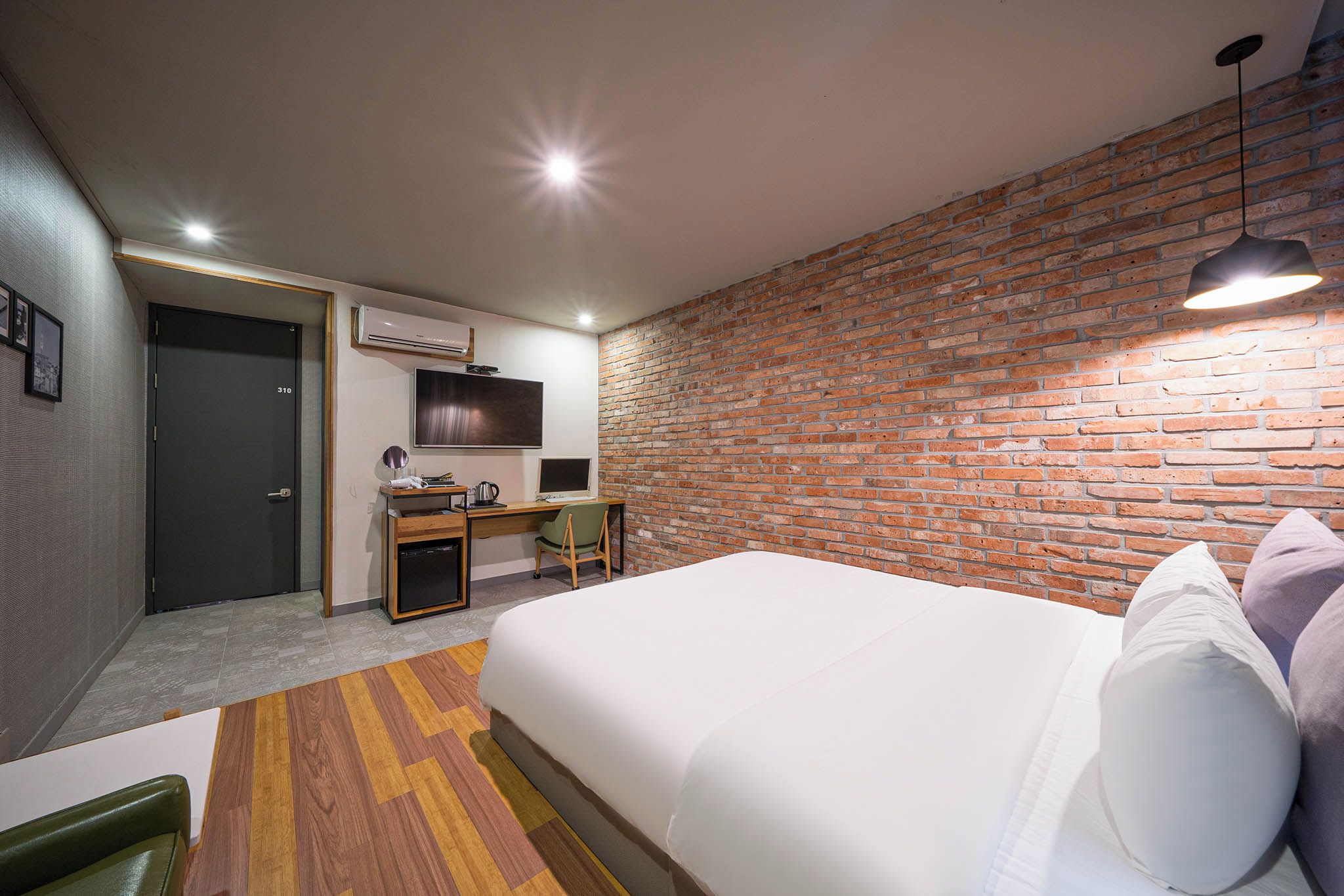 A hotel room with a brick wall, a bed with white bedding, a desk with a computer, and a television.