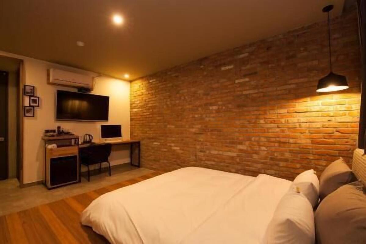 A bedroom with a brick wall, a bed with white bedding, a desk with a computer, and a TV mounted on the wall.