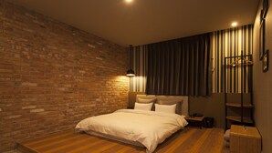 A bedroom with a brick wall, a wooden floor, a bed with white bedding, a nightstand, a lamp, and a hanging coat rack. - Hotel Gray (Changwon)