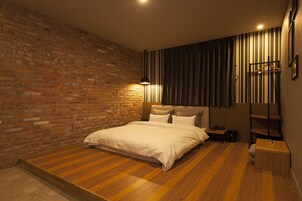 Un dormitorio con pared de ladrillo, suelo de madera, cama con ropa blanca, mesita de noche, lámpara y perchero.