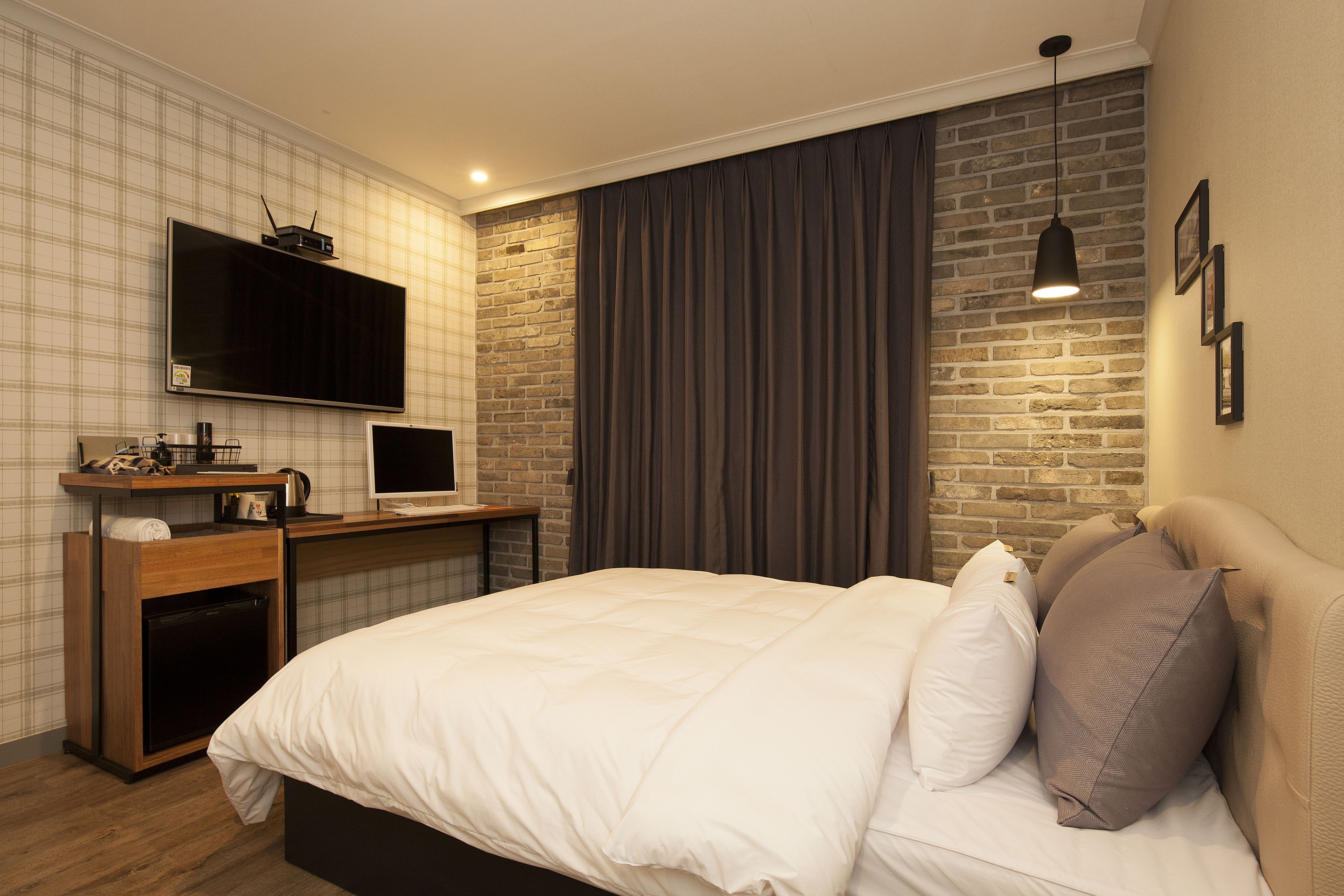 A modern hotel room with a large bed, a flat-screen TV, a desk with a computer, and a brick accent wall.
