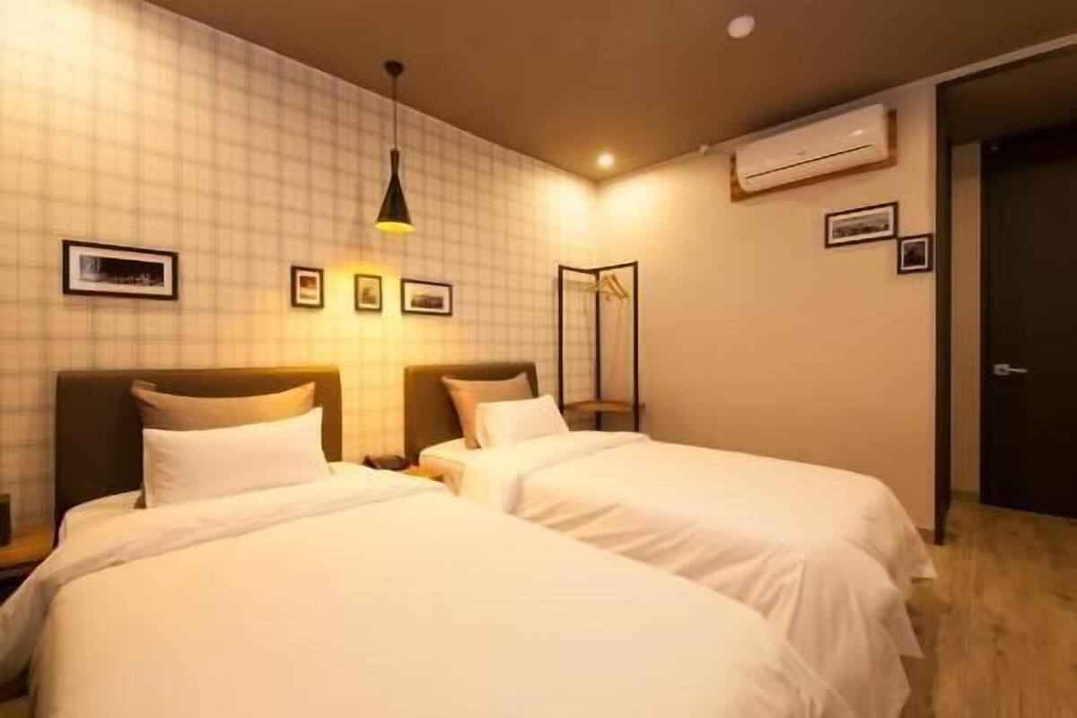 A hotel room with two beds, a hanging light fixture, an air conditioning unit, and framed pictures on the wall.