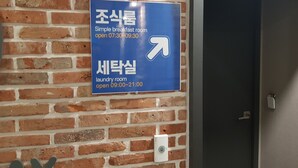 A blue sign on a brick wall indicating a simple breakfast room open from 07:30 to 09:30 and a laundry room open from 09:00 to 21:00. - Hotel Gray (Changwon)