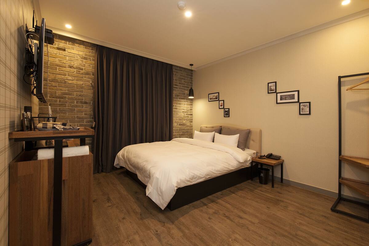 A modern bedroom with a brick accent wall, a large bed, a bedside table with a lamp, and a wooden dresser.