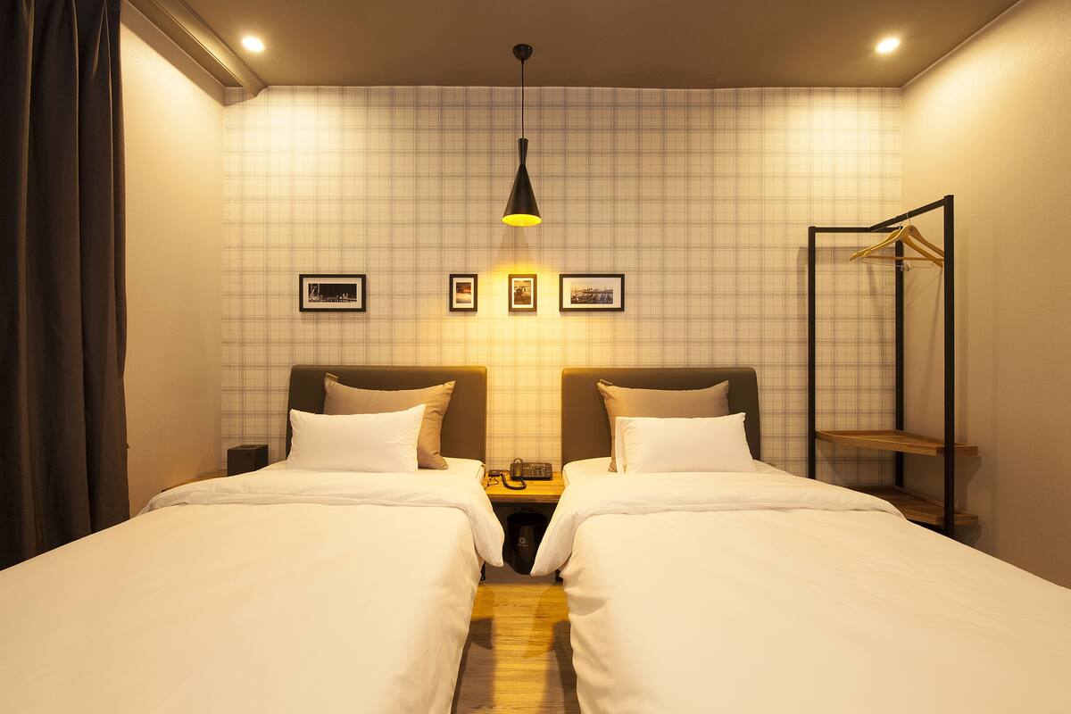 A hotel room with two beds, a bedside table with a lamp, a coat rack, and a wall with framed pictures.